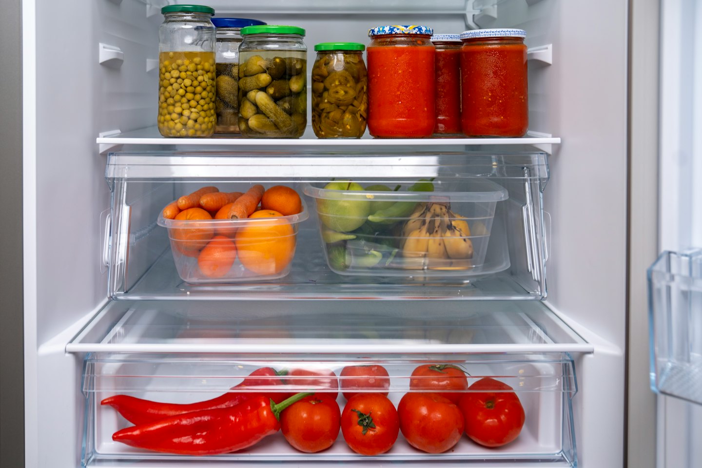 Scaffali del frigorifero organizzati, pieni di frutta fresca, verdura e barattoli di vetro.